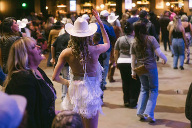 Crowded indoor country dance hall scene with people line dancing, centered on a woman in a white cowboy hat and fringed dress waving her hand.
