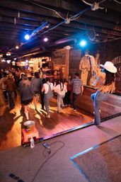 Woman in a white cowboy hat leads a lively line dance on a wooden floor in a dim honky-tonk bar with colorful stage lights and patrons.