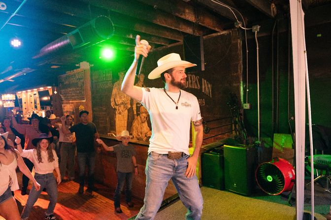 Country singer in a cowboy hat holding a microphone onstage at a lively honky-tonk, crowd line-dancing under green stage lights and rustic wooden interior.