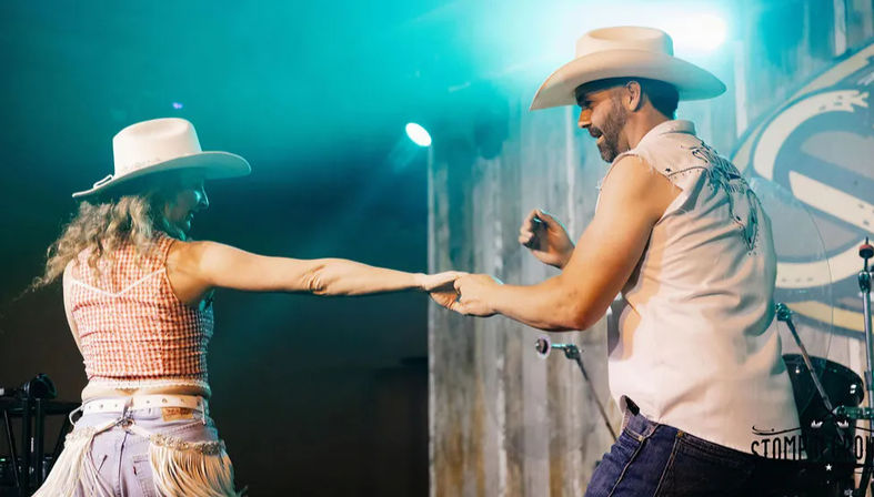 Couple in cowboy hats dancing on a western-themed stage under teal lights, woman in gingham crop top and fringed denim holding hands with smiling man in sleeveless shirt and jeans during a lively country dance.