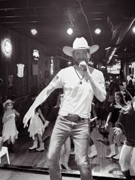 Cowboy-hatted singer with microphone performing on a wooden honky-tonk stage, surrounded by line dancers, neon signs and stage lights.