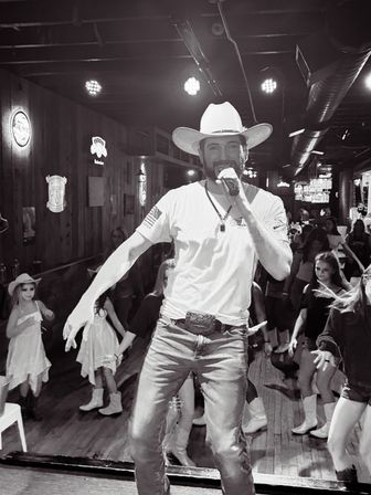 Cowboy-hatted singer with microphone performing on a wooden honky-tonk stage, surrounded by line dancers, neon signs and stage lights.