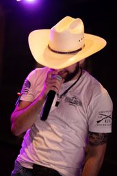 Country singer in a white cowboy hat and casual t-shirt singing into a microphone on stage under purple lights, showing a tattooed arm and belt buckle.