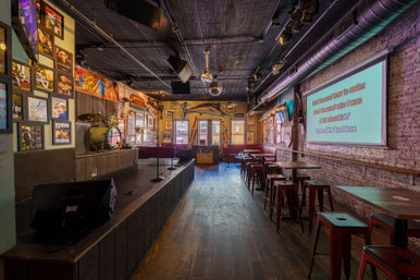 Cozy live-music bar interior with a small stage, drum kit and microphones, disco ball and framed posters, plus a karaoke screen projecting lyrics above red metal stools and wooden tables.