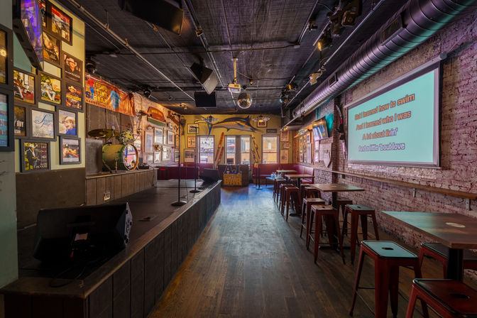 Cozy live-music bar interior with a small stage, drum kit and microphones, disco ball and framed posters, plus a karaoke screen projecting lyrics above red metal stools and wooden tables.