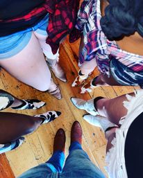 Overhead shot of a circle of people wearing cowboy boots, denim shorts and tied plaid shirts on a rustic wooden floor — playful western-style outfit photo.