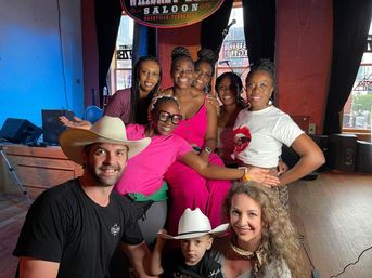 Smiling group of adults and a child posing on a small wooden stage in a Nashville, TN live-music saloon, with cowboy hats, colorful outfits, microphone and speakers visible.