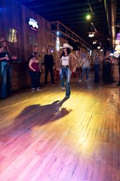 Cowgirl in a white cowboy hat and fringe jacket line-dancing in a spotlight on a wide wooden dance floor inside a rustic country bar, patrons watching along the walls.