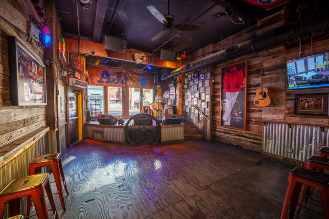 Rustic live-music bar interior with empty wooden dance floor, small raised stage with drum kit and microphones, guitars and framed memorabilia on wood-paneled walls, neon signs and metal bar stools.
