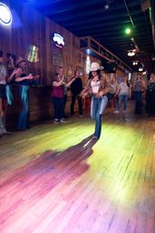 Woman in a white cowboy hat line-dancing on a wooden dance floor in a lively country bar with colorful stage lights and a cheering crowd by rustic wood-paneled walls.