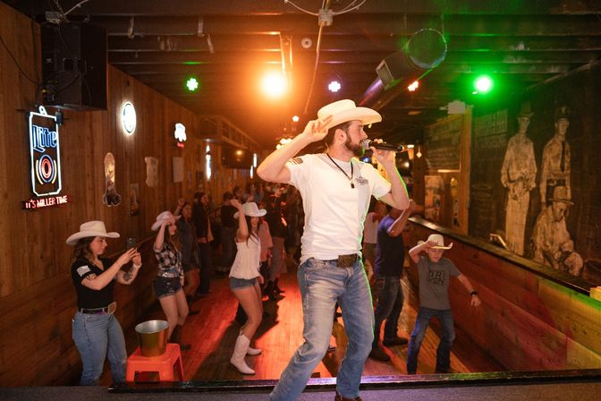 Energetic country bar scene: singer in a cowboy hat on stage with a microphone while patrons in cowboy hats line-dance on a wood-paneled dance floor under warm stage lights.