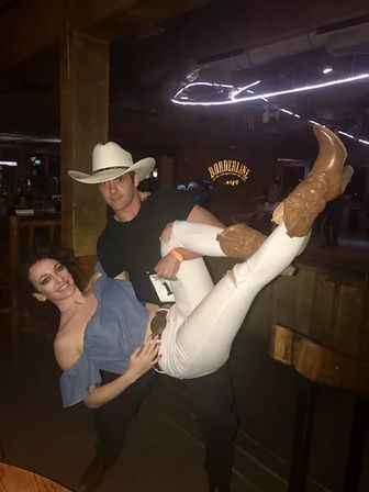 Playful pose of a man in a white cowboy hat lifting a woman in an off-shoulder blue top, white ripped jeans and brown cowboy boots inside a dim, wooden country bar with neon lights.