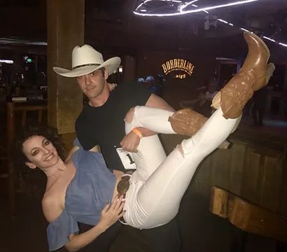 Playful pose of a man in a white cowboy hat lifting a woman in an off-shoulder blue top, white ripped jeans and brown cowboy boots inside a dim, wooden country bar with neon lights.