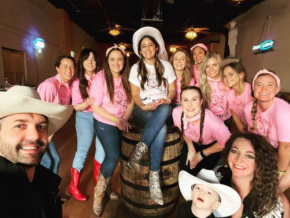 Bachelorette party group photo in a rustic western bar — women in pink shirts, cowboy hats and boots gathered around a wooden barrel, with a man and child in cowboy hats.