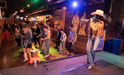 Toe‑tapping country bar scene: female singer in a cowboy hat and fringe jacket on stage leading a lively crowd line dancing on a wooden floor under colorful lights with a rustic bar and mural behind them.