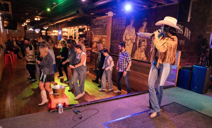 Toe‑tapping country bar scene: female singer in a cowboy hat and fringe jacket on stage leading a lively crowd line dancing on a wooden floor under colorful lights with a rustic bar and mural behind them.