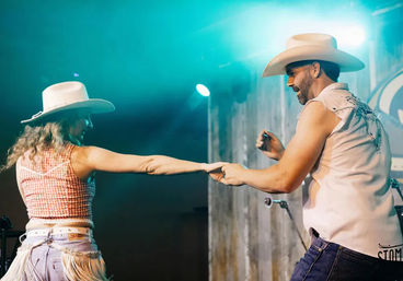 Two country dancers in cowboy hats and western clothing hold hands mid-spin on a teal-lit stage, smiling and wearing fringe jeans and sleeveless shirts.