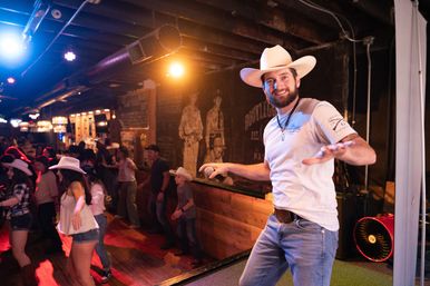 Smiling male emcee in a white cowboy hat and jeans holding a microphone on stage as a lively crowd line-dances under warm lights in a rustic country bar.
