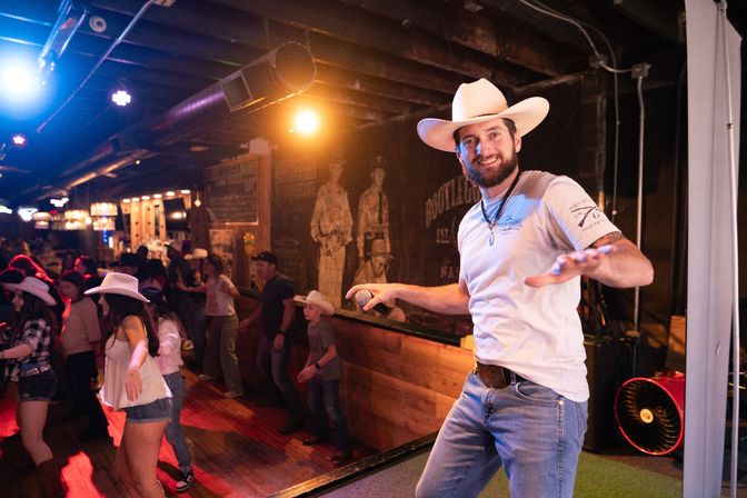 Smiling male emcee in a white cowboy hat and jeans holding a microphone on stage as a lively crowd line-dances under warm lights in a rustic country bar.