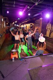 Crowd dancing on a wooden dance floor in a lively country bar, performer in a white cowboy hat on stage under purple lights