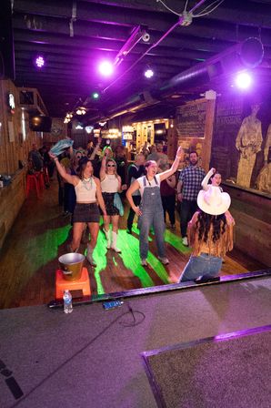 Crowd dancing on a wooden dance floor in a lively country bar, performer in a white cowboy hat on stage under purple lights