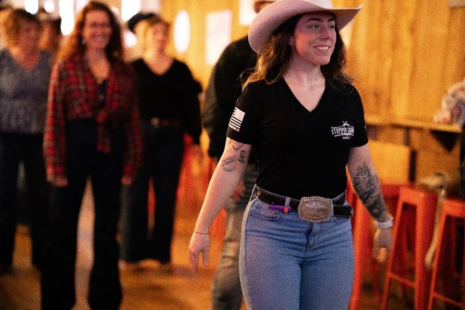 Smiling woman in a cowboy hat, black t-shirt and jeans with a large belt buckle leading a line of dancers at a warm‑lit country line‑dancing night in a barn‑style dance hall.