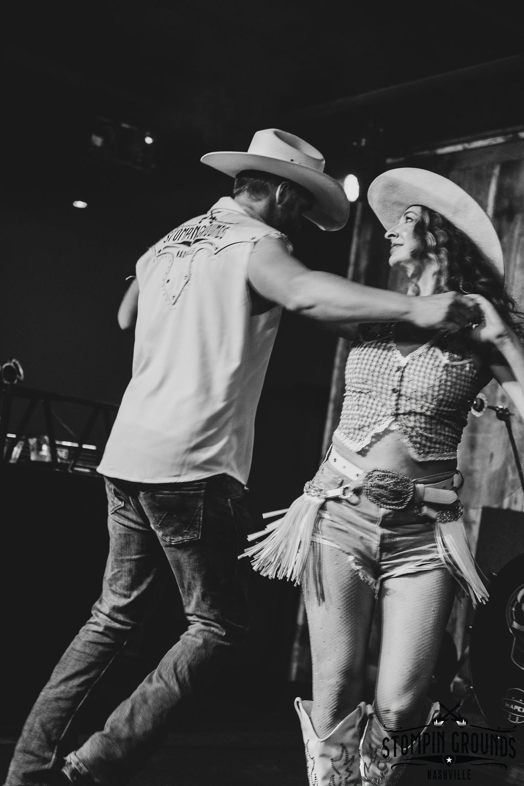 Energetic black-and-white shot of two country dancers mid-twirl, wearing cowboy hats, fringed belt, denim and boots on a dimly lit Nashville stage.