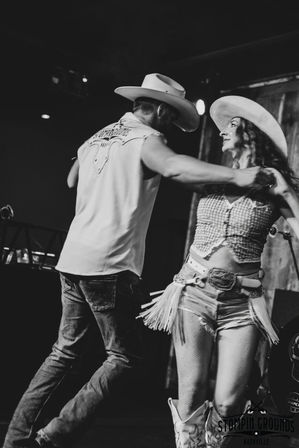Energetic black-and-white shot of two country dancers mid-twirl, wearing cowboy hats, fringed belt, denim and boots on a dimly lit Nashville stage.