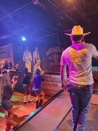 Man in a cowboy hat on a small stage leading a group of dancers on a wooden floor at a lively Nashville country bar, colorful stage lights and a rustic mural in the background.