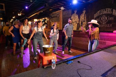 Adults boot-scootin' and line-dancing to live country music in a rustic wooden bar, led by a cowgirl singer in a cowboy hat onstage under purple lights.