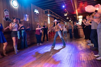 Country bar scene with a wooden dance floor as a smiling woman in a cowgirl hat, fringe jacket and jeans leads line dancing while a crowd of patrons in boots and casual clothes clap and cheer.
