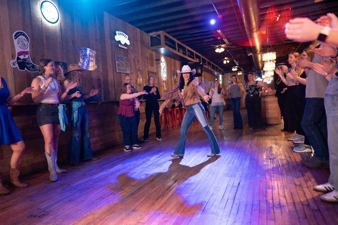 Country bar scene with a wooden dance floor as a smiling woman in a cowgirl hat, fringe jacket and jeans leads line dancing while a crowd of patrons in boots and casual clothes clap and cheer.