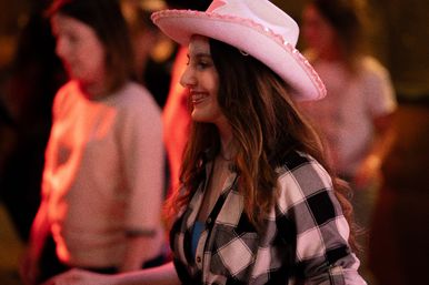 Smiling woman wearing a pink cowboy hat and plaid shirt at a lively country dance night with warm red lighting