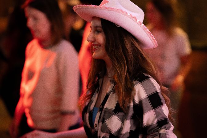 Smiling woman wearing a pink cowboy hat and plaid shirt at a lively country dance night with warm red lighting