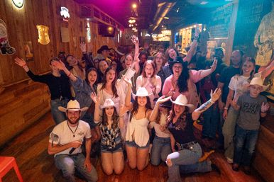 Group of smiling people cheering in a rustic country bar, many wearing cowboy hats with hands raised amid wooden walls and neon signs — lively country-nightlife scene.