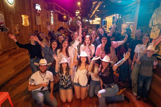Group of smiling people cheering in a rustic country bar, many wearing cowboy hats with hands raised amid wooden walls and neon signs — lively country-nightlife scene.