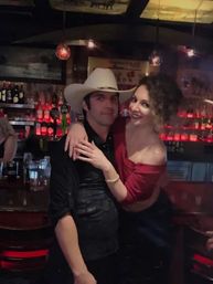 Smiling couple at a dim saloon-style bar — man wearing a white cowboy hat and black shirt and a woman in an off-shoulder red top hugging him, red backlit bottles and pendant lights on the bar shelves behind them.