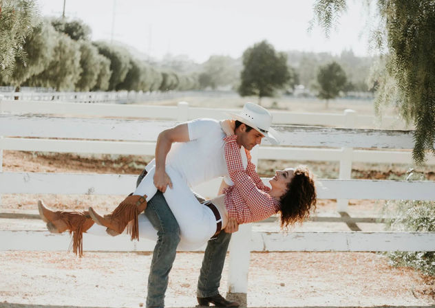 Playful country couple — man in a cowboy hat dips a woman in a gingham shirt and fringe boots by a white ranch fence on a sunny farm