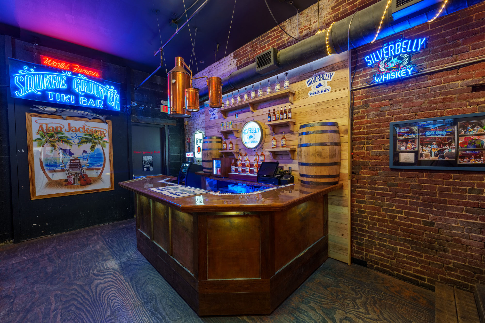 Vibrant tiki-style bar interior with neon signage, wooden L-shaped bar, whiskey bottle display and barrel against exposed brick under copper pendant lights.