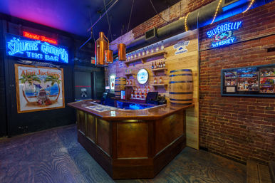 Vibrant tiki-style bar interior with neon signage, wooden L-shaped bar, whiskey bottle display and barrel against exposed brick under copper pendant lights.
