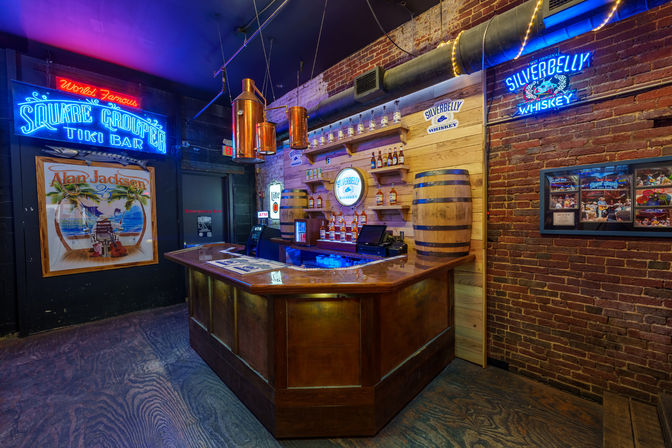 Vibrant tiki-style bar interior with neon signage, wooden L-shaped bar, whiskey bottle display and barrel against exposed brick under copper pendant lights.