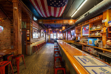 Cozy rustic American bar interior with a long polished wooden counter, rows of red metal stools, hanging U.S. flag, exposed brick walls, neon signs, and stocked liquor shelves.
