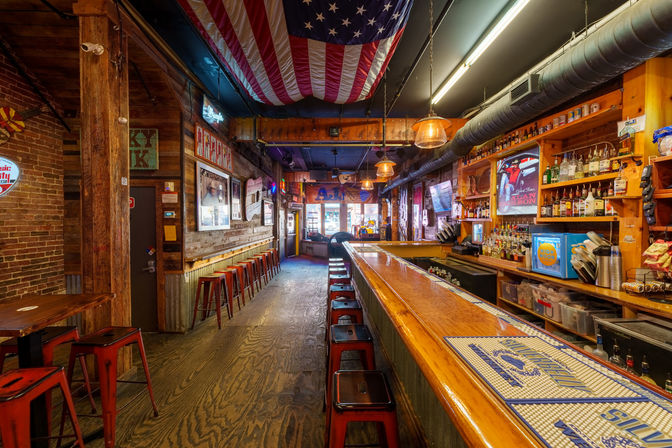 Cozy rustic American bar interior with a long polished wooden counter, rows of red metal stools, hanging U.S. flag, exposed brick walls, neon signs, and stocked liquor shelves.