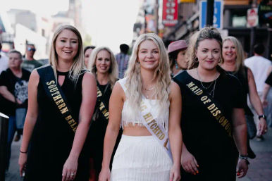 Group of women on a busy downtown street celebrating a bachelorette: the bride-to-be in a white fringe outfit wearing a sash reading Getting Hitched, flanked by friends in black dresses with sashes like Southern Belle and Maid of Dishonor, all smiling.