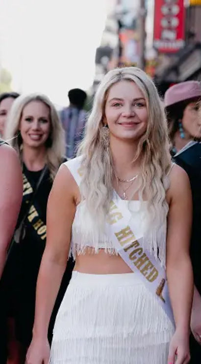 Group of women on a busy downtown street celebrating a bachelorette: the bride-to-be in a white fringe outfit wearing a sash reading Getting Hitched, flanked by friends in black dresses with sashes like Southern Belle and Maid of Dishonor, all smiling.