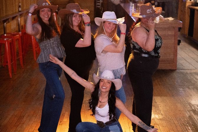 Group of five women in cowboy hats striking a playful country-western pose on a wooden dance hall floor with red bar stools in the background