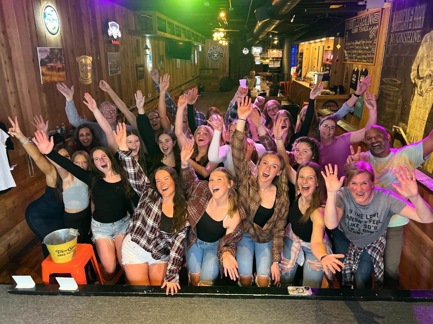 Group photo of friends partying in a rustic wood-paneled bar — about two dozen people, mostly young women in casual plaid and denim, cheering with hands raised under colorful lights.