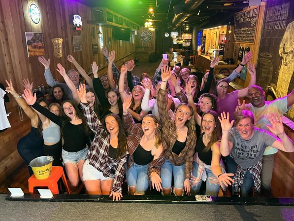 Group photo of friends partying in a rustic wood-paneled bar — about two dozen people, mostly young women in casual plaid and denim, cheering with hands raised under colorful lights.