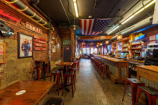 Rustic honky-tonk country bar interior with exposed brick walls, wooden floors and long wooden bar lined with red metal stools, American flag hanging from the ceiling, framed country music posters, string lights and neon signs.