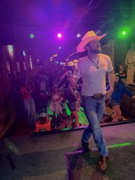 Live country singer in a cowboy hat on stage with a microphone as a lively crowd line-dances under colorful neon lights in a rustic bar